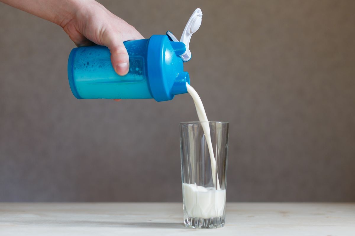 Ready milk whey cocktail protein drink is poured from a shaker into a glass Cup. Sports drink for a set of pure muscle mass. Hand holding plastic shaker with protein shake and glass on table